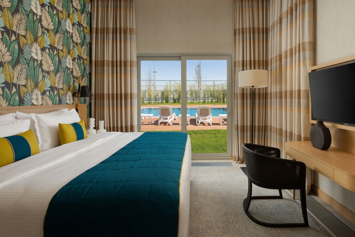 A hotel room with a bed, a desk with a TV, a chair, and a view of an outdoor pool area.