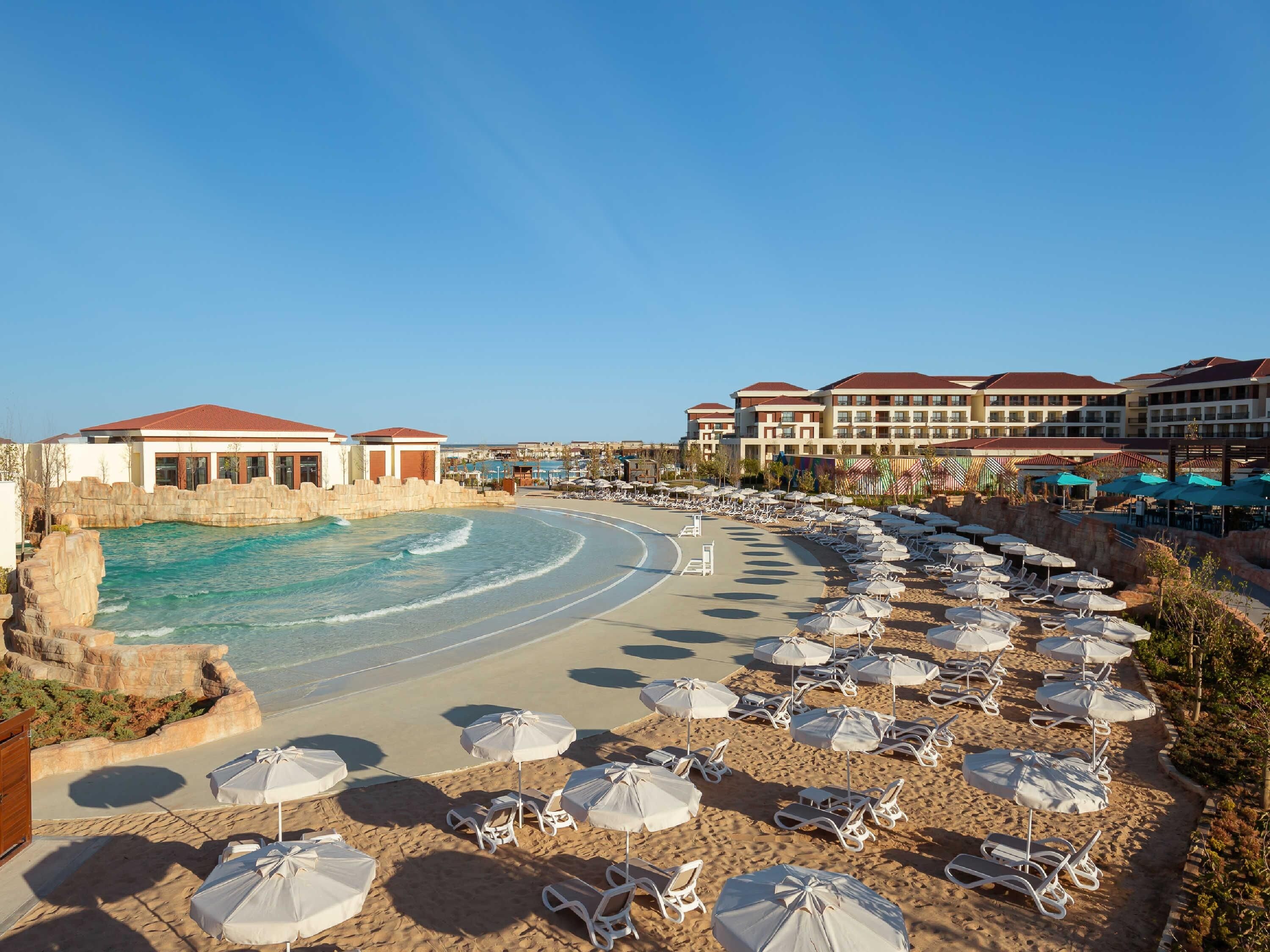 Et resort med pool, strand med liggestole og parasoller, samt hotelbygninger i baggrunden.