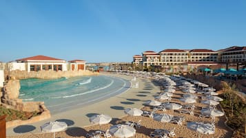 Et resort med pool, strand med liggestole og parasoller, samt hotelbygninger i baggrunden.