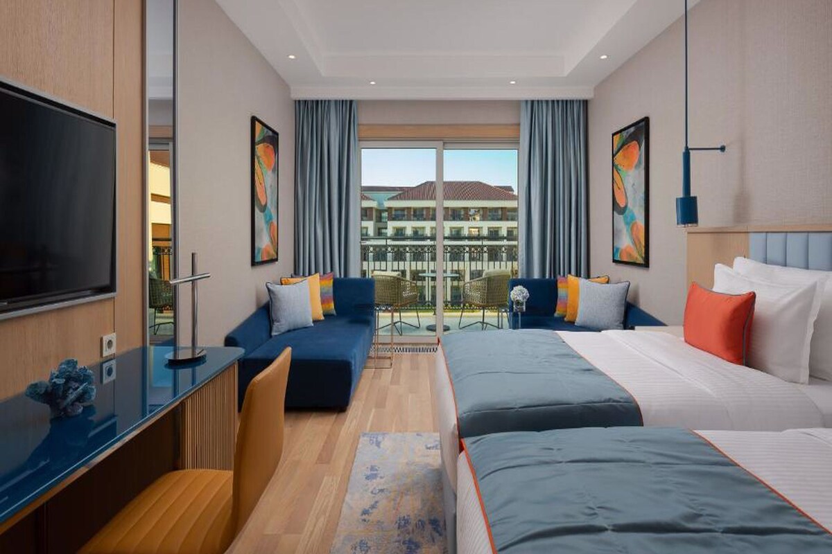 A modern hotel room with a large bed, a desk, and a sofa. There is a balcony with a view of buildings.