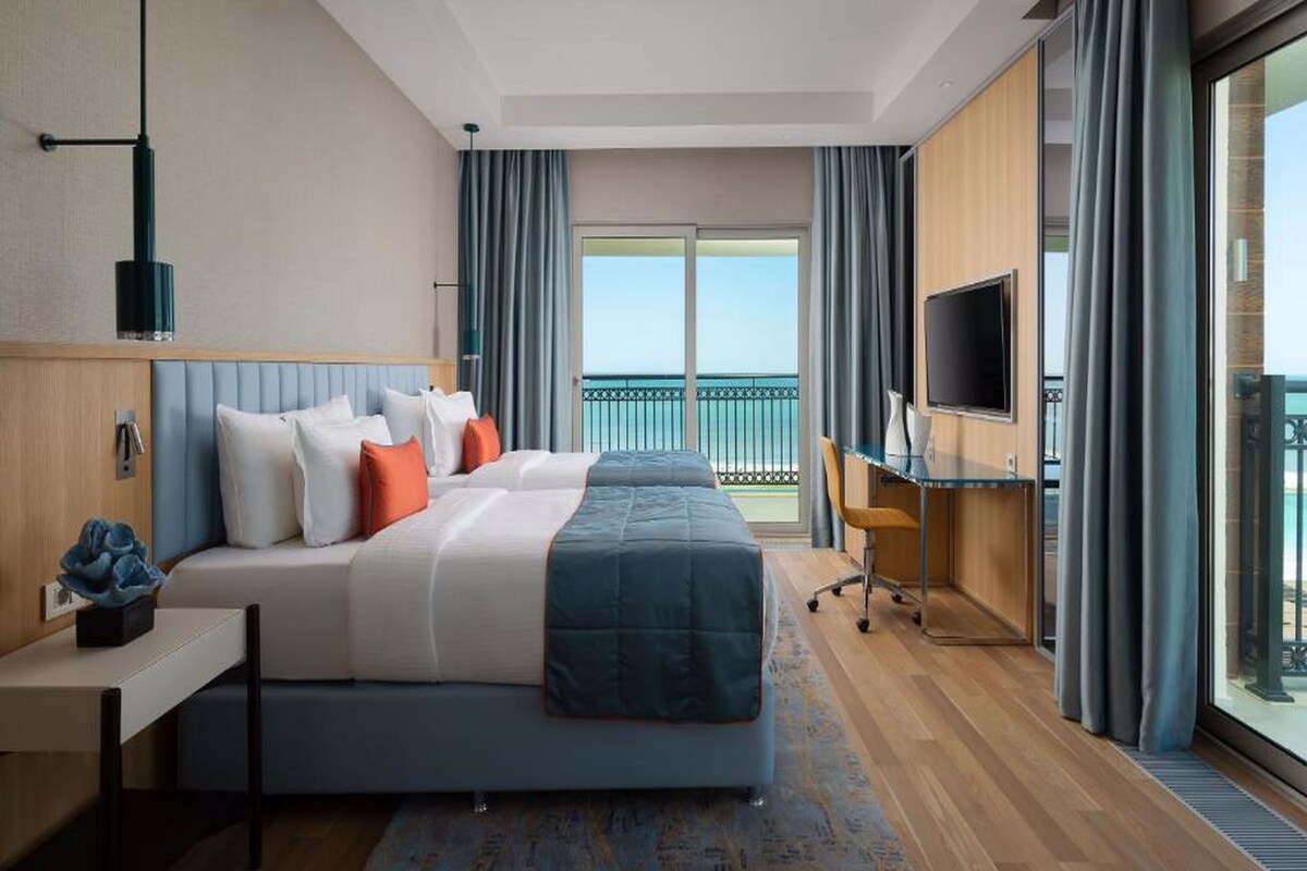 A hotel room with a large bed, a desk, a TV, and a balcony with a view of the sea.