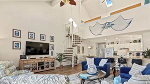 A spacious living room with a large TV, comfortable seating, a staircase, and a kitchen in the background.