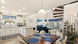A modern kitchen with white cabinets, a dining area with a wooden table and blue placemats, and a spiral staircase with a railing.