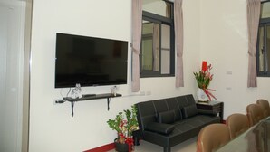 A living room with a flat-screen TV mounted on the wall, a black sofa, a glass dining table with chairs, and a potted plant. - Eliza Inn (Magong)