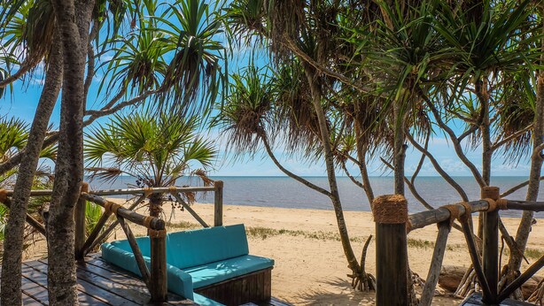 A wooden deck with a blue cushioned seating area, surrounded by palm trees and overlooking a sandy beach. - Saadani Safari Lodge - bush, river, beach (Saadani)