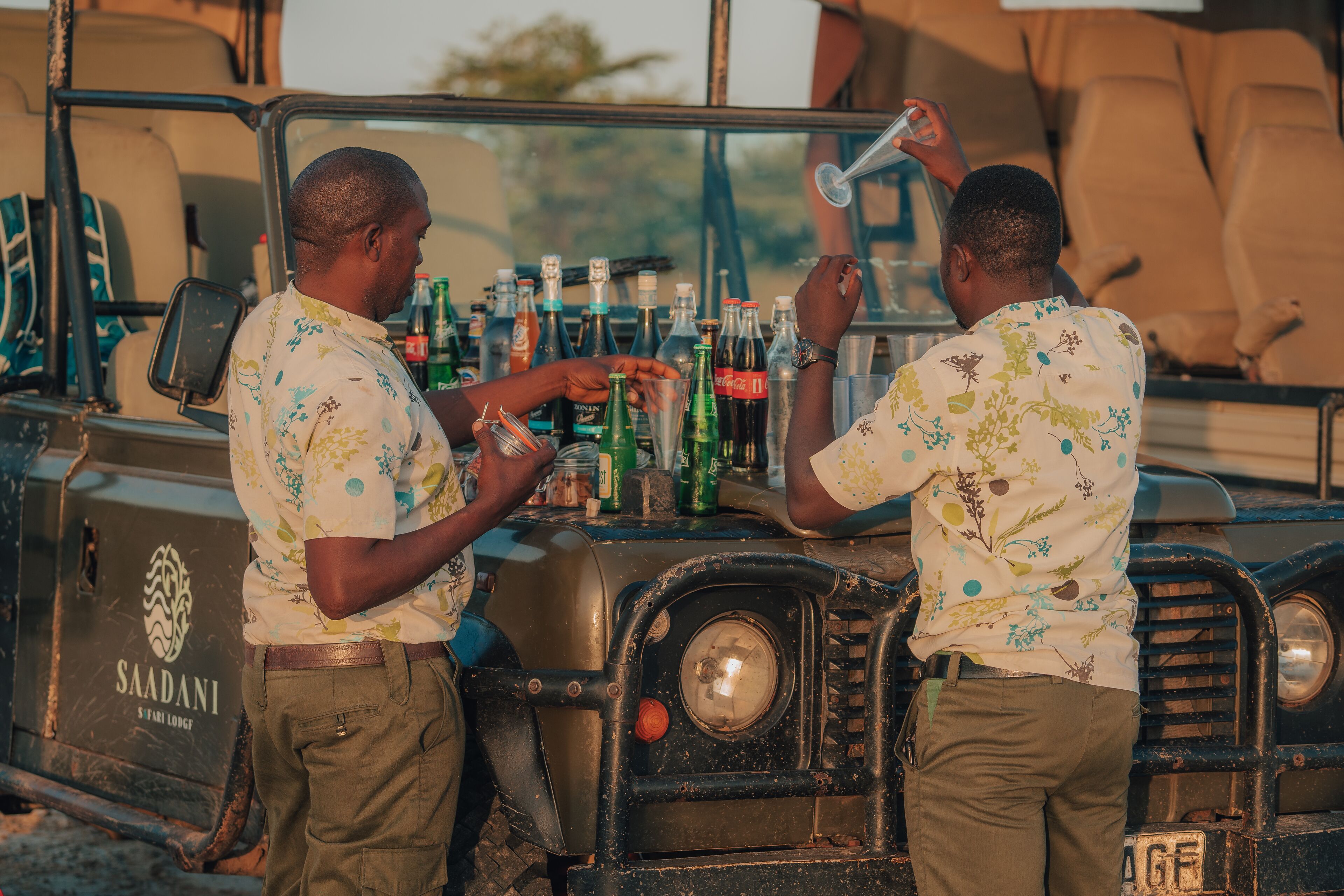 Two men standing by a vehicle with a bar area, bottles, and a sign that reads "SAADANI S VARI LODGE".