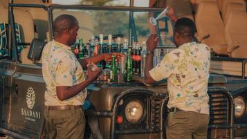 Two men standing by a vehicle with a bar area, bottles, and a sign that reads "SAADANI S VARI LODGE".