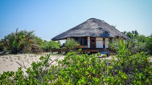A beach house with a thatched roof, surrounded by greenery and sand. - Saadani Safari Lodge - bush, river, beach (Saadani)