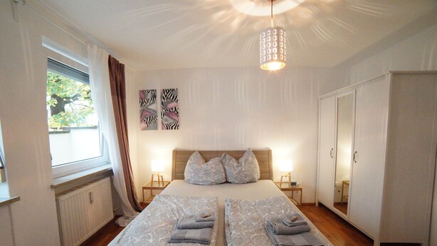 Uma cama de casal bem arrumada com almofadas e toalhas dobradas, uma mesa de cabeceira com abajur, uma janela com cortinas e um guarda-roupa com espelho.
