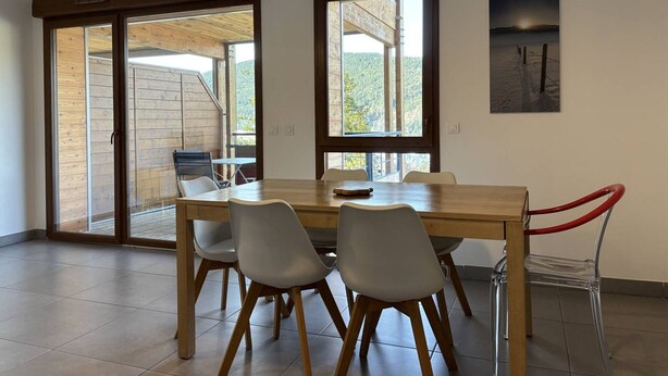 A dining area with a wooden table and chairs, large windows, and a view of a wooden deck outside. - Cozy 2-Bedroom Apartment with Balcony & Free Parking in Villard-de-Lans (Villard-de-Lans)