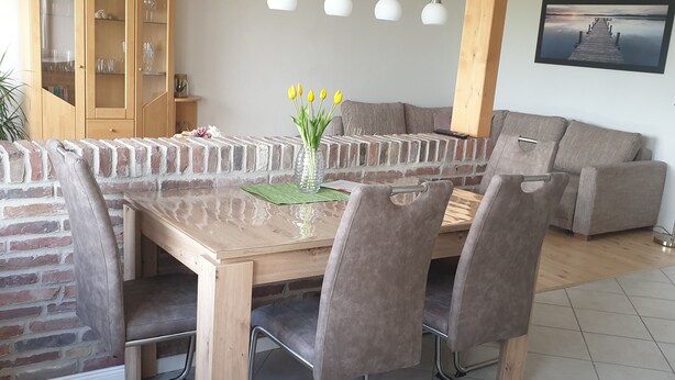 A dining area with a brick partition, a wooden table, grey upholstered chairs, and a grey sofa. - Vacation apartment "Auf der Warf" (Wittmund)