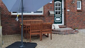 A patio area with a table and chairs, a grey umbrella, and a brick building with a green door in the background. - Vacation apartment "Auf der Warf" (Wittmund)