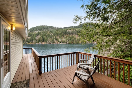 A wooden deck with a railing, overlooking a lake and forested hills.