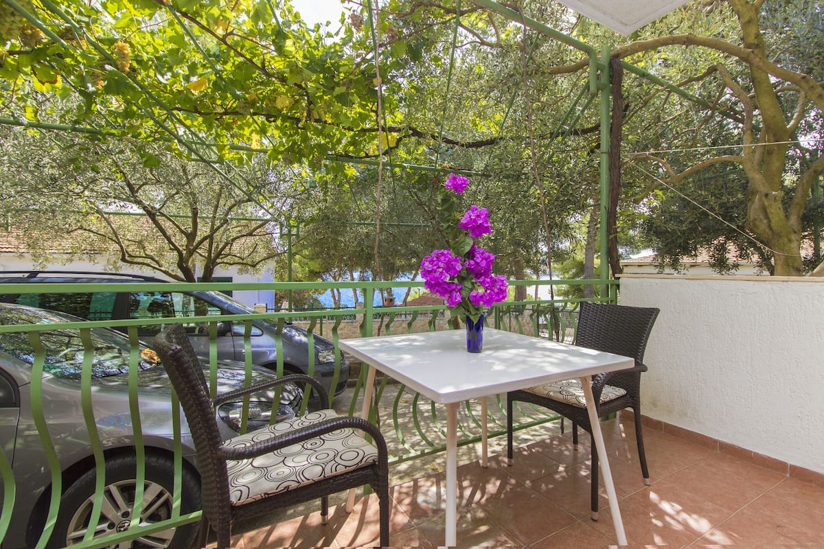 Un patio con mesa y sillas, un coche aparcado cerca y un árbol con flores moradas al fondo.