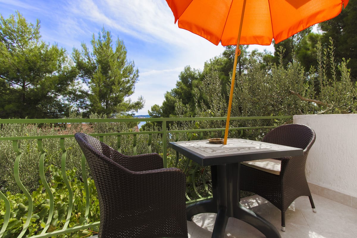 Un patio al aire libre con sillas de mimbre y una mesa, barandal verde y vistas a árboles y un cuerpo de agua.