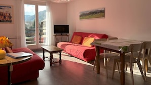 Una sala de estar con un sofá rojo, una mesa de madera, un televisor sobre un mueble bajo y un balcón con vistas a montañas.