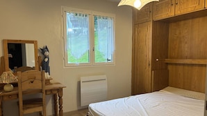 Un dormitorio con un escritorio de madera, una silla, una cama, una ventana con cortinas y un armario de madera.