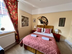 Ein Hotelzimmer mit Bett, zwei Nachttischen, einem Fenster mit Vorhängen, einer Heizung und Wandbildern.