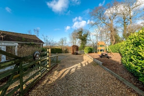 A gravel path leading to a wooden gate, a hay bale, and a play structure in a rural setting.