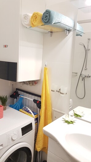 Casa de banho com máquina de lavar roupa, pia, chuveiro e toalhas numa estante.