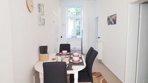 Un coin repas avec une table blanche dressée pour quatre personnes, des chaises noires et une fenêtre ornée d’une pancarte indiquant « BIENVENUE ».