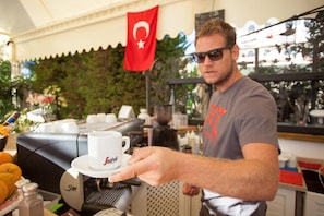 A person holding a cup with a logo, a coffee machine, and a Turkish flag in the background. - Mitos Apartments (İçmeler)