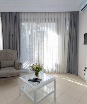 A modern living room with a sofa, a glass coffee table with remotes, a vase with flowers, and a TV on the wall. - Mitos Apartments (İçmeler)