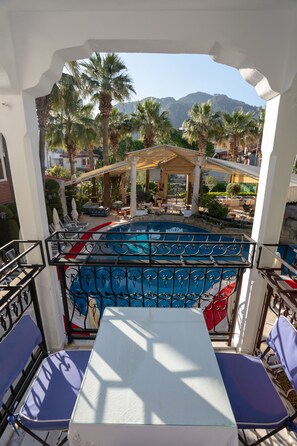 A balcony with a view of a pool, palm trees, and a mountain in the background. - Mitos Apartments (İçmeler)