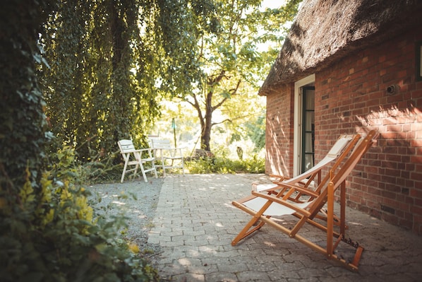 A brick building with a thatched roof, a wooden easel, and a white chair in a garden setting.