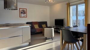 Una cocina moderna con una isla central, un comedor con mesa y sillas, un sofá con cojines, un televisor y un amplio ventanal con cortinas.