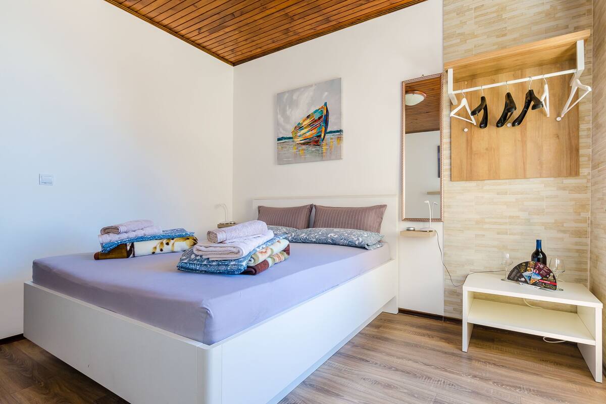 A neatly made bed with folded towels, a bedside table with a bottle and glasses, and a coat rack with hangers.