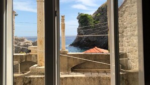 A view through a window of a coastal area with stone structures and a clear sky. - Rooms City Central Old Town (Dubrovnik)