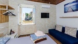 A room with a wooden ceiling, a TV mounted on the wall, a window with an open door, a sofa with patterned cushions, and a bed with a blue and white blanket. - Rooms City Central Old Town (Dubrovnik)