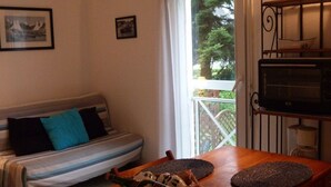 Uma sala de estar acolhedora, com um sofá, uma mesa de jantar com jogos americanos, uma cesta de petiscos e vista para o verde lá fora.