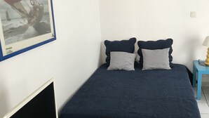 Quarto com cama, escrivaninha com computador, quadro na parede e pequena mesa com abajur.