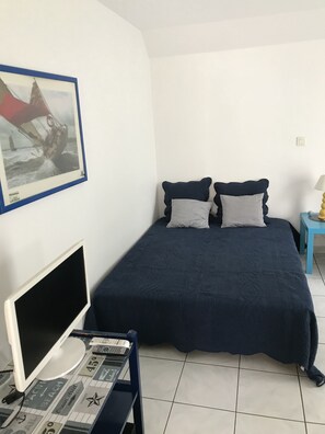 A bedroom with a bed, a desk with a computer, a framed picture on the wall, and a small table with a lamp.