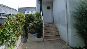 Uma escadaria de concreto estreita que leva a uma porta, cercada por vegetação e uma parede branca.