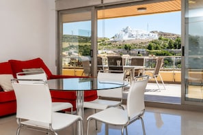 A modern living room with a glass table and white chairs, a red sofa, and a balcony with a view of a white building on a hill.