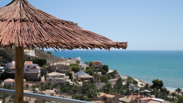 Ein Balkon mit einem Reetdach-Sonnenschirm mit Blick auf eine Küstenstadt, mit Häusern und dem Meer.