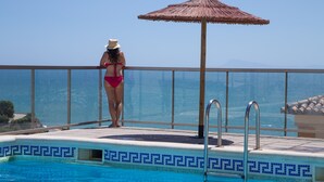 Eine Frau in einem Bikini steht an einem Pool mit einem Reetdachschirm und blickt auf das klare, blaue Meer.