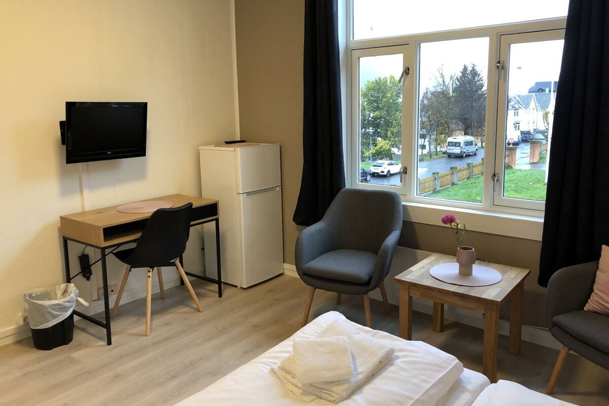 Habitación de hotel con cama, escritorio con computadora, silla, pequeña mesa con un florero y ventana con vista a la calle.