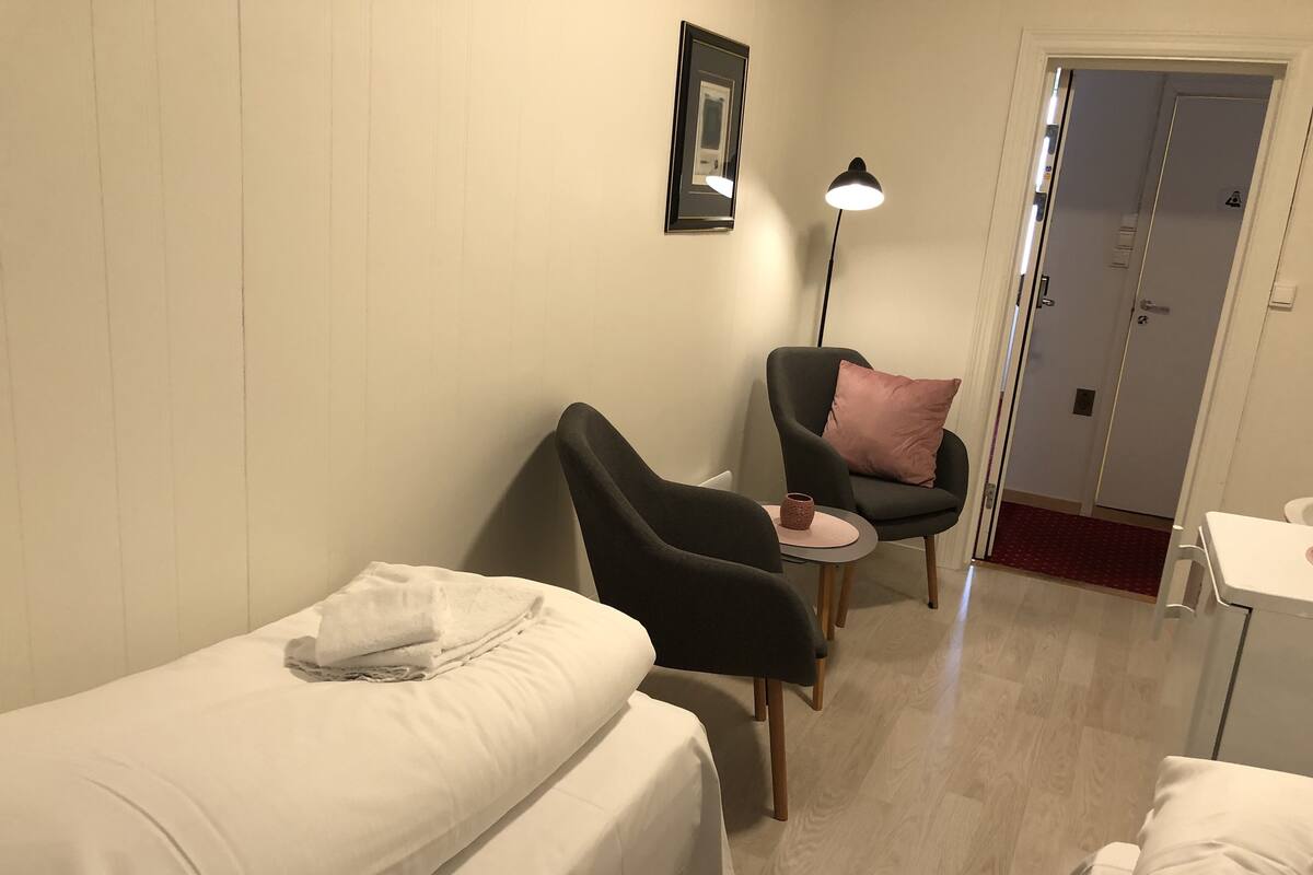 Una habitación pequeña y ordenada con dos camas individuales, una silla con un cojín rosa, una mesita con una taza y una lámpara de pie.