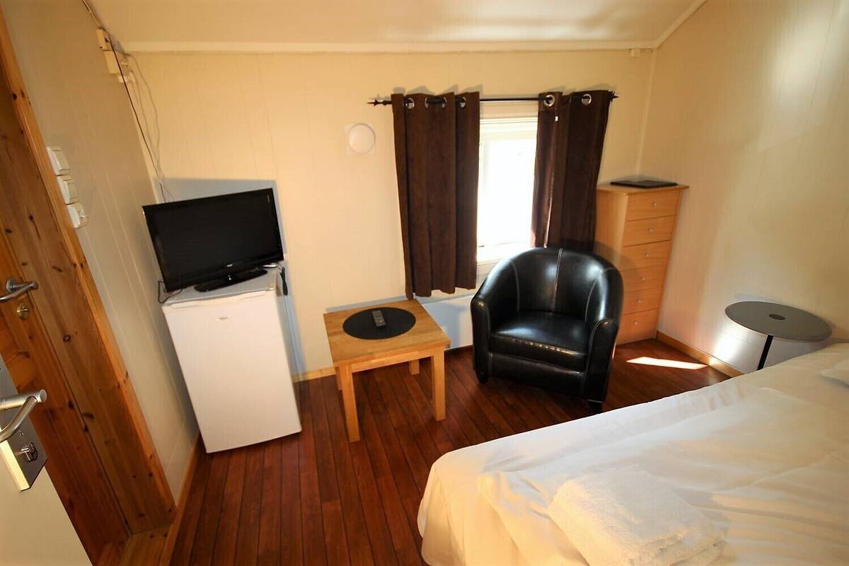 Una habitación de hotel funcional con cama, escritorio con silla, televisor sobre un mini refrigerador y armario.