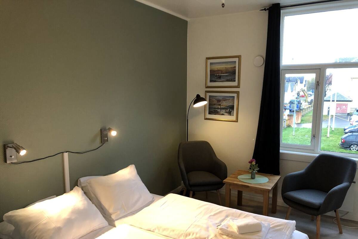 Un cuarto de hotel con una cama, dos sillas, una mesita con un florero y dos cuadros en la pared.