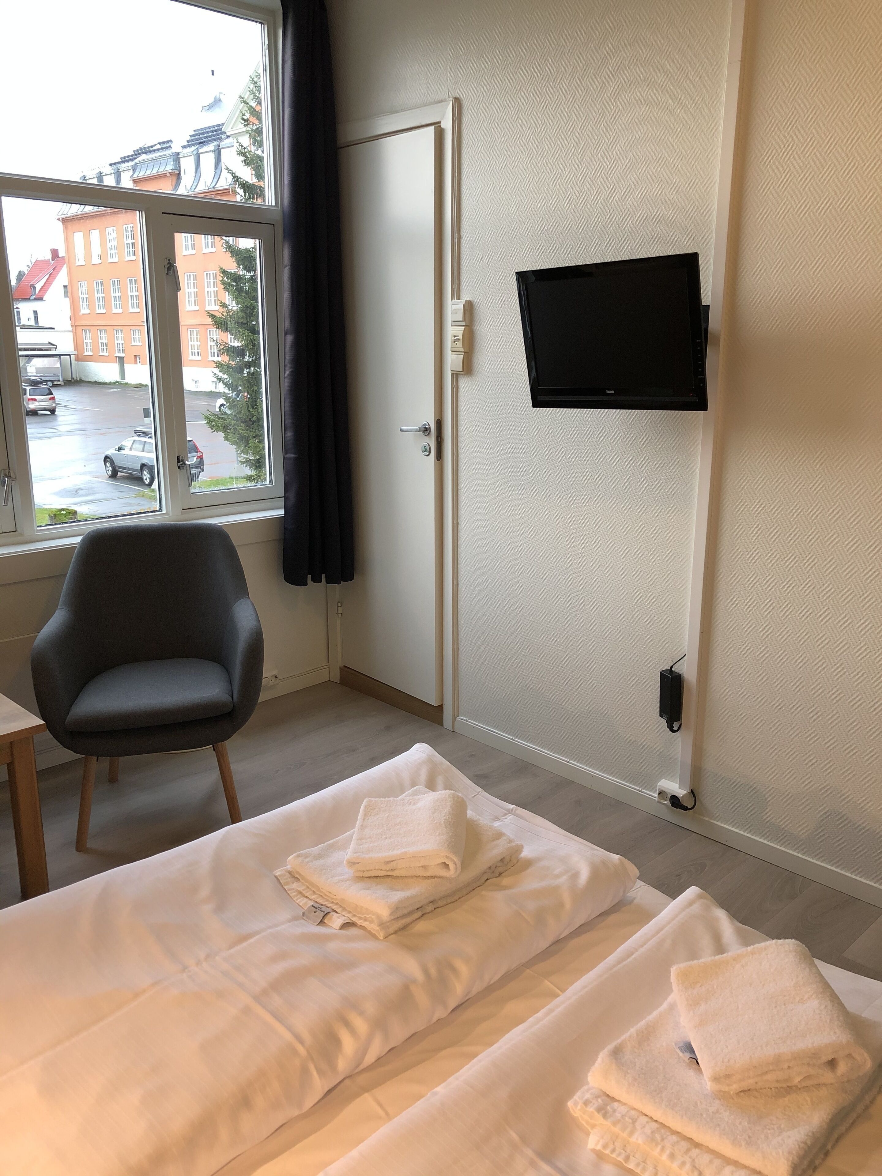 Una habitación de hotel con cama, una silla, un televisor montado en la pared y una ventana con vistas a edificios y coches aparcados.