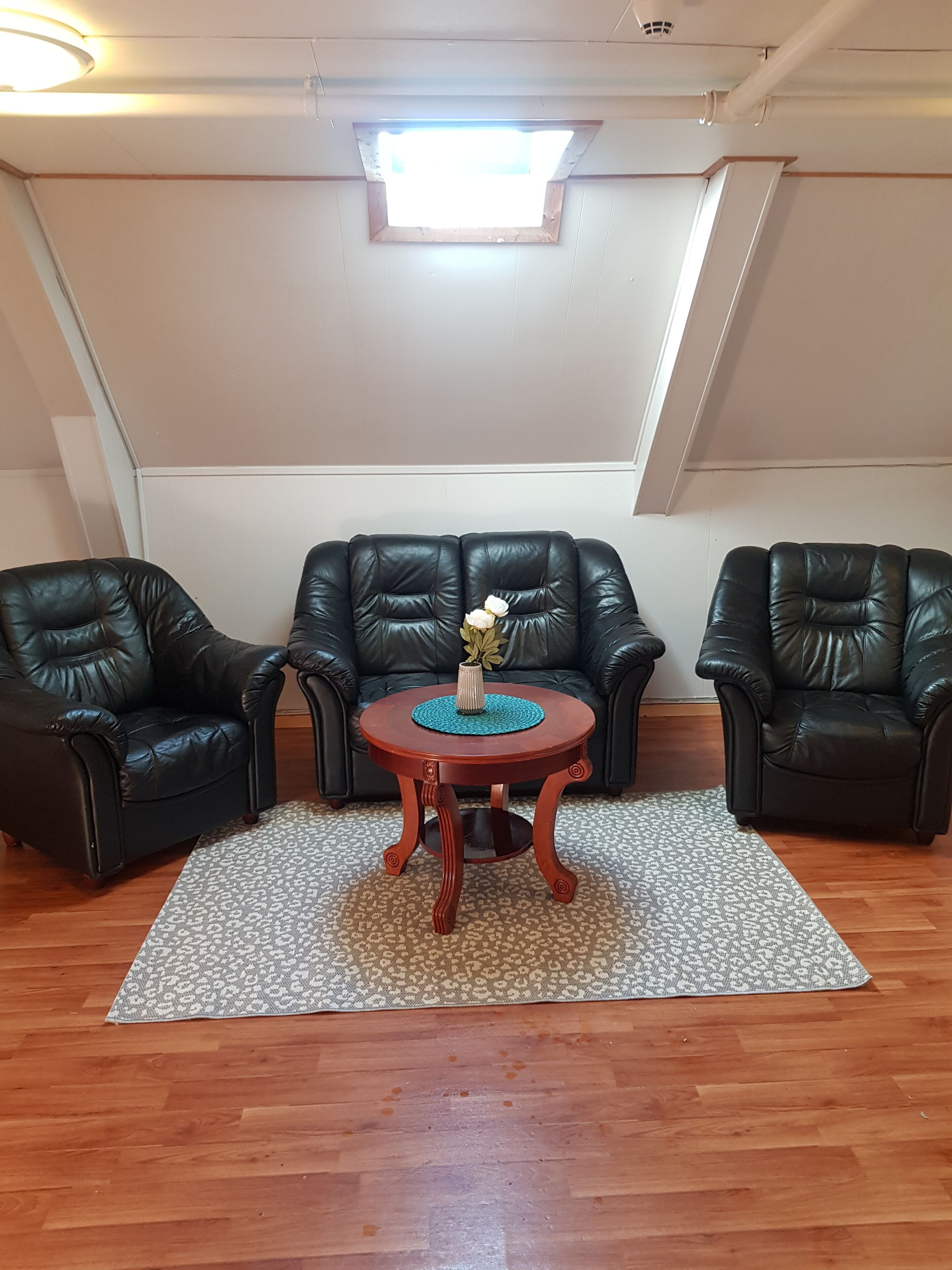 A room with a wooden floor, a patterned rug, two black leather chairs, a small wooden table with a vase of flowers, and a skylight above.