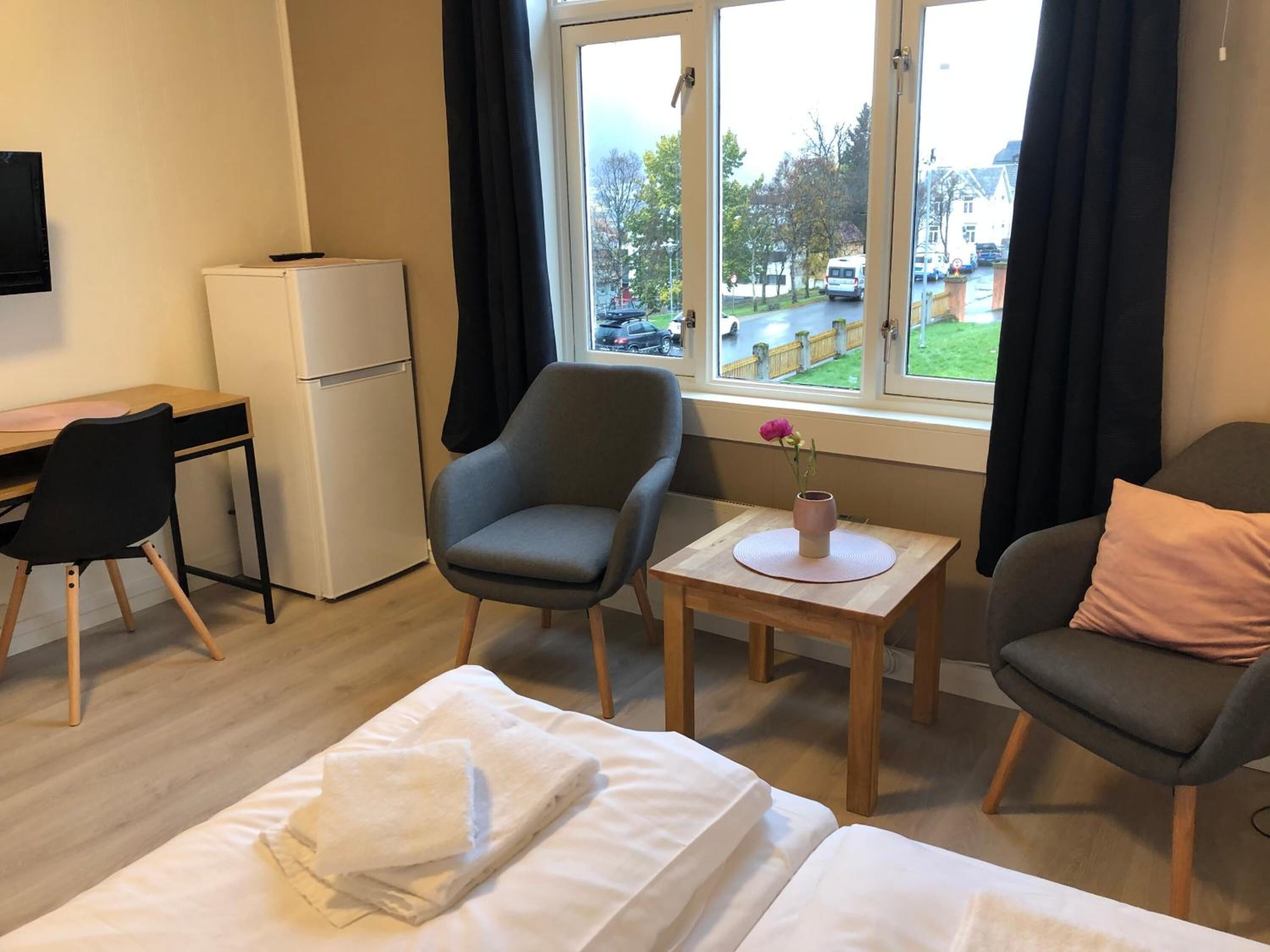Habitación de hotel con cama, dos sillas, una mesita con un florero, un escritorio con computadora y un refrigerador.
