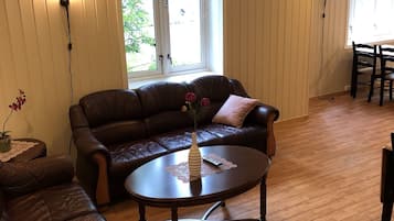 Una sala de estar con un sofá de cuero marrón, una mesa redonda de centro con un jarrón y un control remoto, piso de madera y una ventana con cortinas.