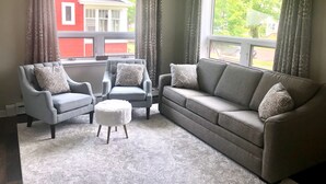 Un soggiorno con un divano grigio, due poltrone, un poggiapiedi bianco e cuscini decorativi. Sono presenti due finestre con tende, una ventola a soffitto e vista su un edificio rosso.