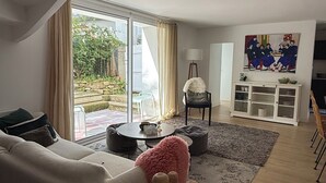 A modern living room with a sofa, a round coffee table, a pink pouf, and a wooden side table. There is a dining area with a chair and a cabinet with dishes. A painting is on the wall, and a window offers a view of the outdoors.